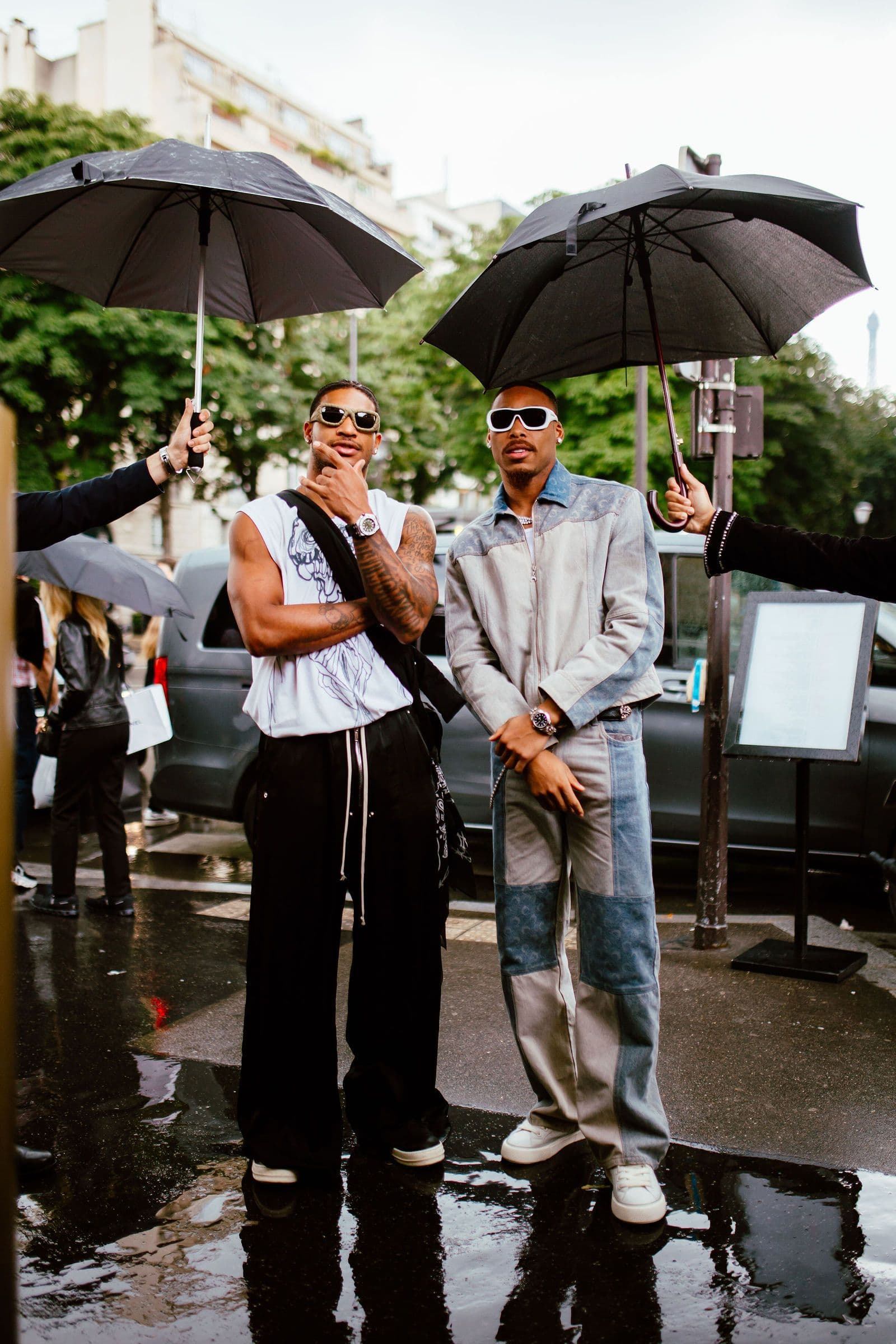 Two men stand under umbrellas on a wet street, looking at the camera